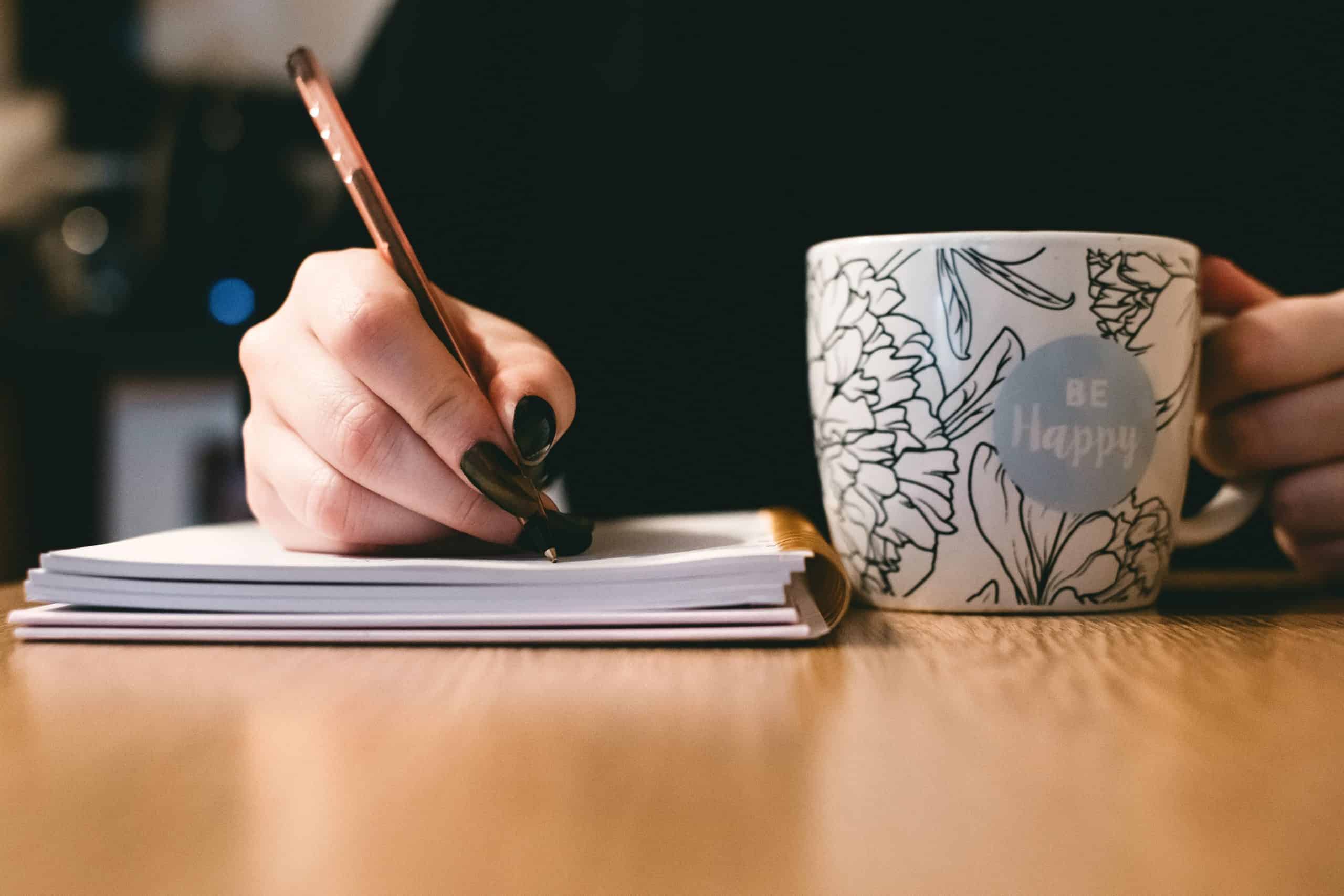 A woman with black pointed nails holds a coffee mug that says, "be happy" and writes in a journal