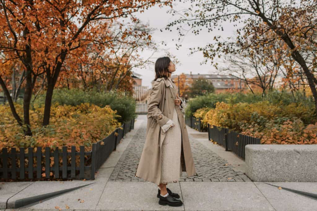 A woman stands on the sidewalk near a park and looks up to the sky.