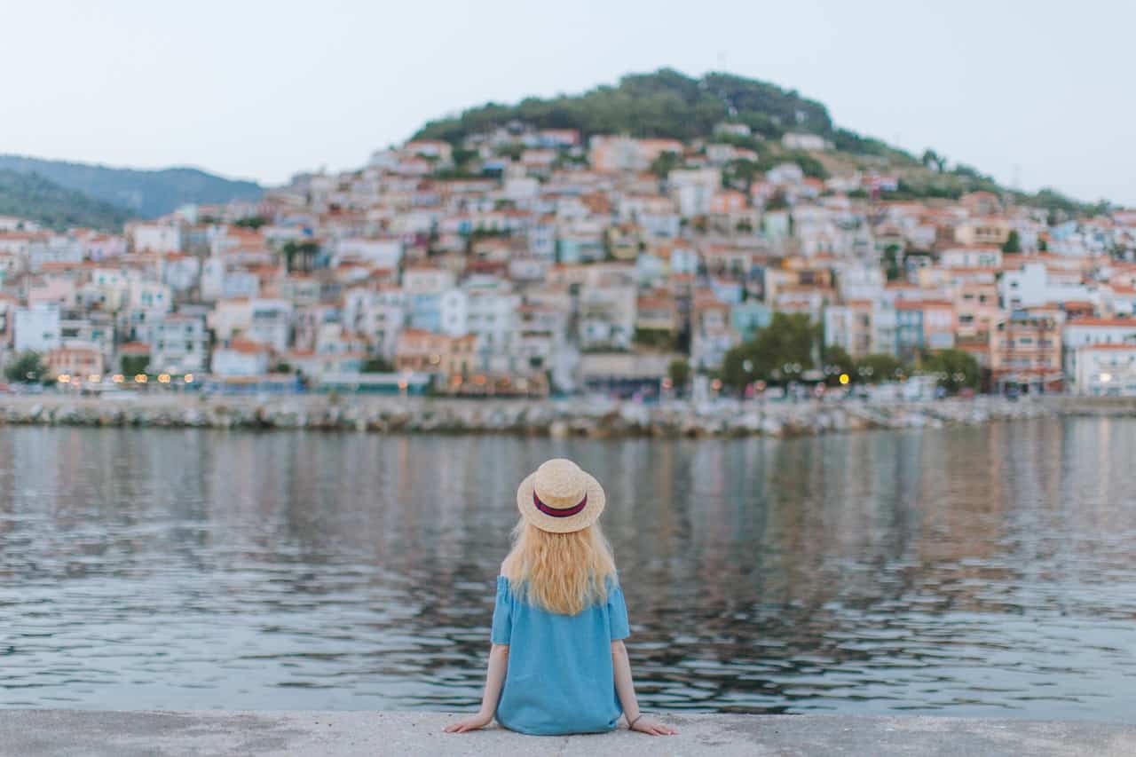 a woman sits near water bordering a village built on a hill