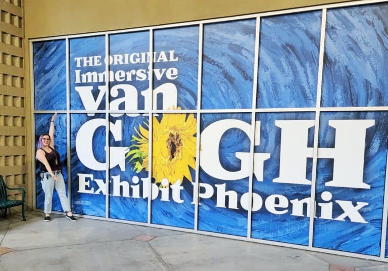 A woman poses in front of a sign that says, "The original immersive van gogh exhibit phoenix"