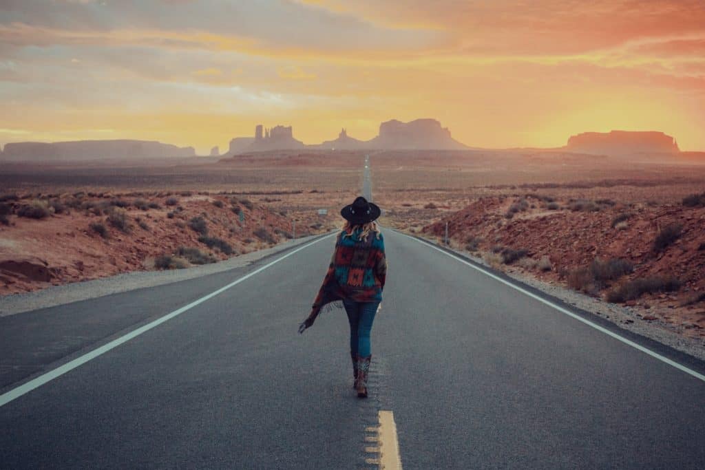 A blond woman in a multi-colored patched poncho and a black hat stands in the middle of the road in the American Southwestern desert