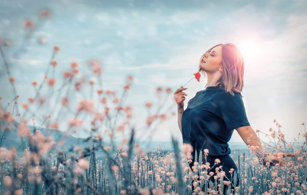 A woman holds a poppy flower up to her face while standing in the middle of a field of flowers.