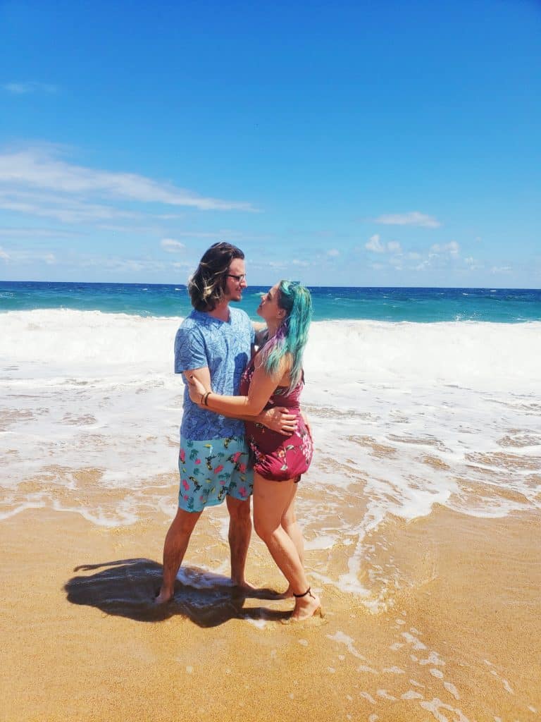 A couple stands on a sandy beach with their feet in the water, starring into each other's eyes.