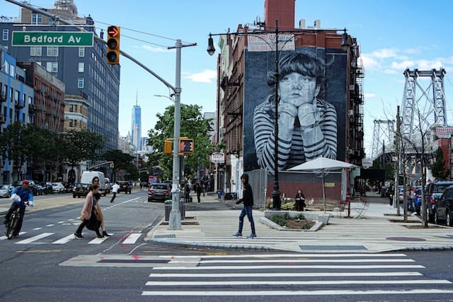 The cross roads at Bedford Av featuring a wall mural showcasing a girl with her head on her hands.