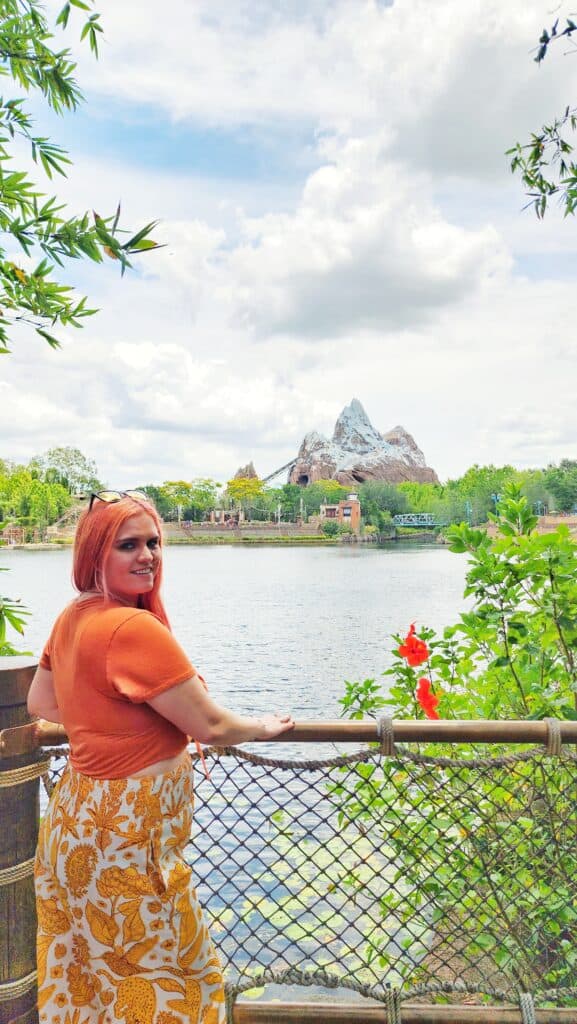 A woman stands against a railing in front of a body of water between her and a faraway mountain.