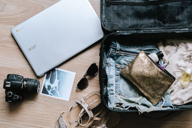 A suitcase lays open on the floor with a laptop, camera, sandals, and sunglasses beside it.