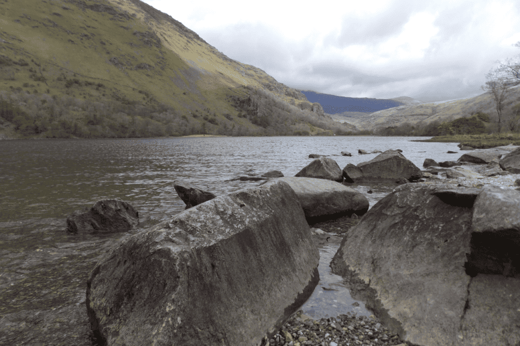 A cloudy ski hovers over a still lake with rock boulders in the forefront