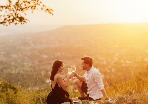 A couple sits on top of a hill at sunset and have a picnic