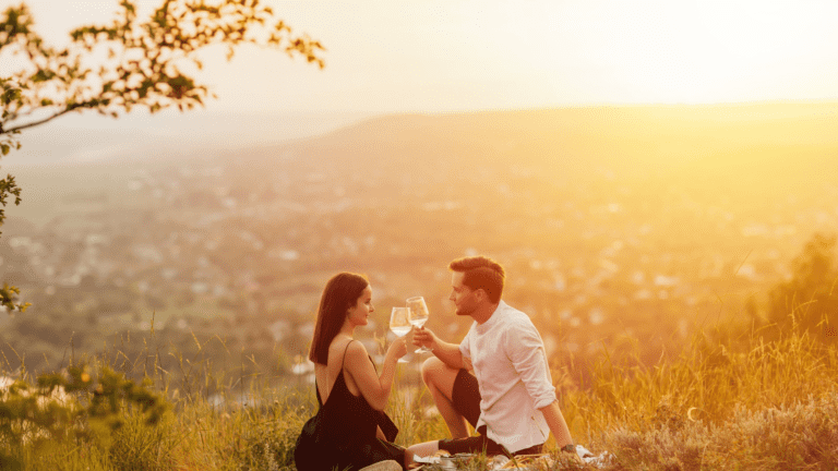 A couple sits on top of a hill at sunset and have a picnic