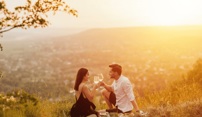A couple sits on top of a hill at sunset and have a picnic