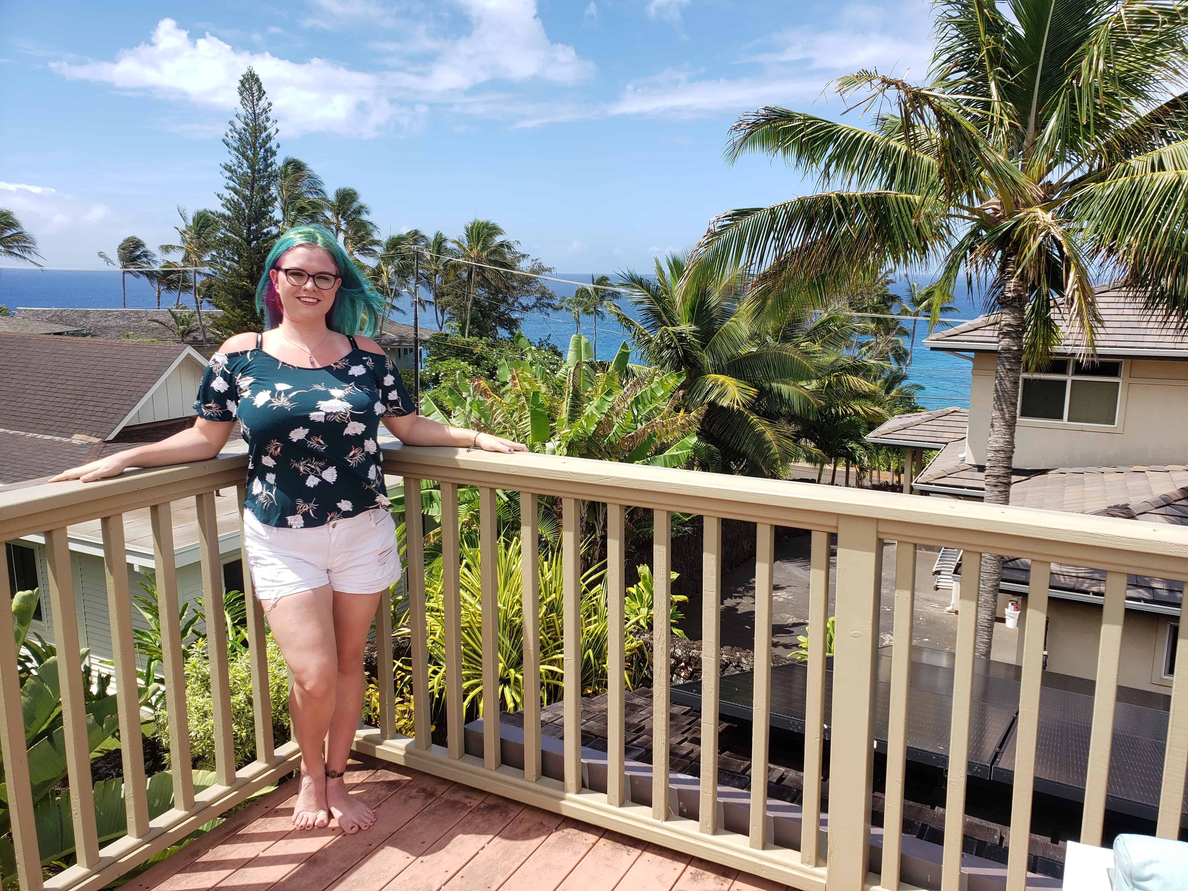 A woman with green and purple hair wearing a green floral shirt and white shorts stands against a balcony. The view behind her is residential with palm trees and the ocean in the distance behind her.