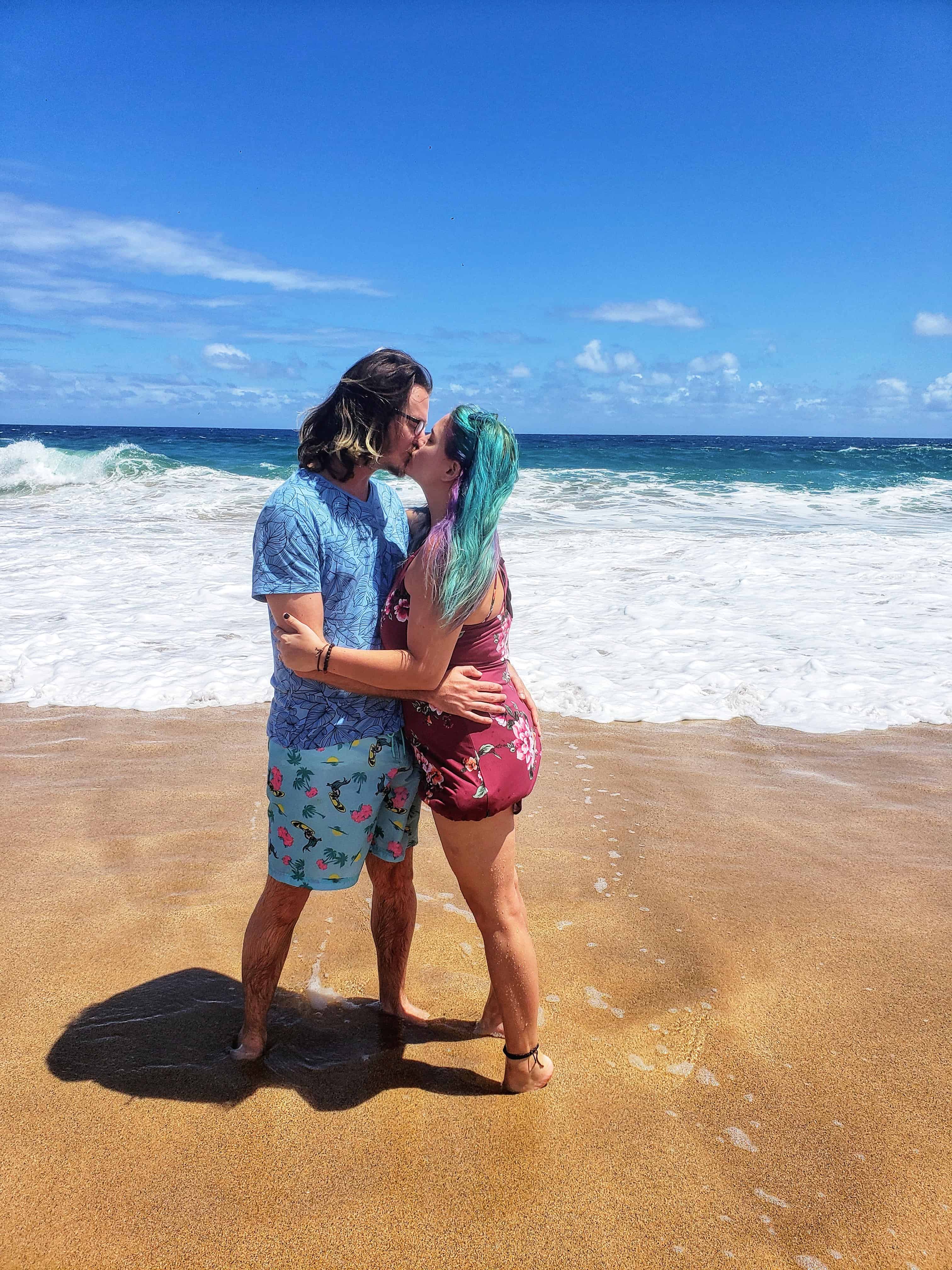 A man in blue swim trunks and a blue t-shirt and woman in a burgundy dress kiss on a white sandy beach.