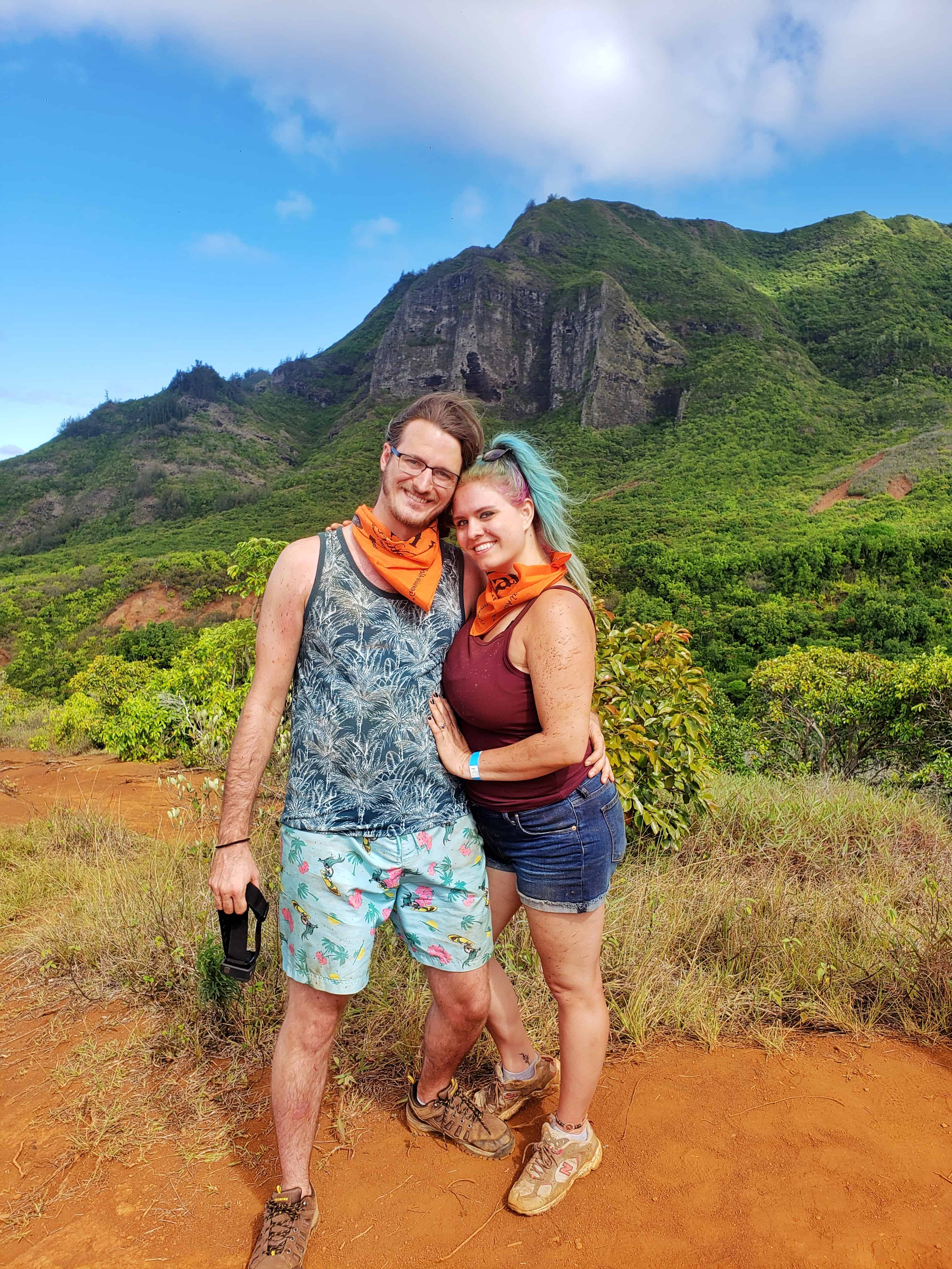 A man and a woman pose next to each other for a picture. The backdrop is of a tall, tropical mountain.