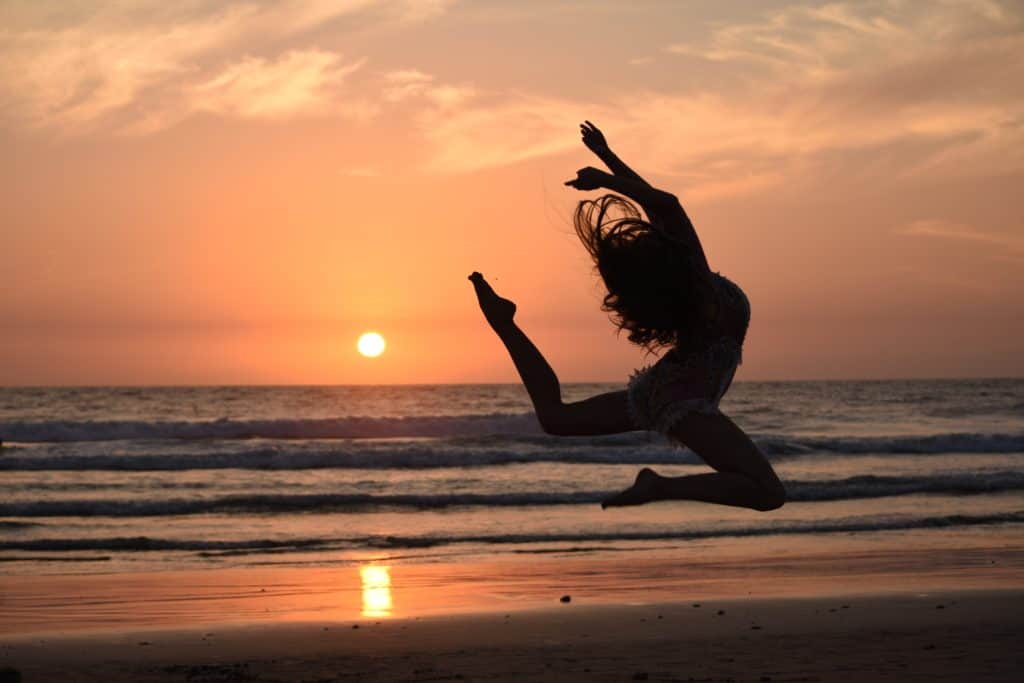 A woman leaps through the air on a beach at sunset.
