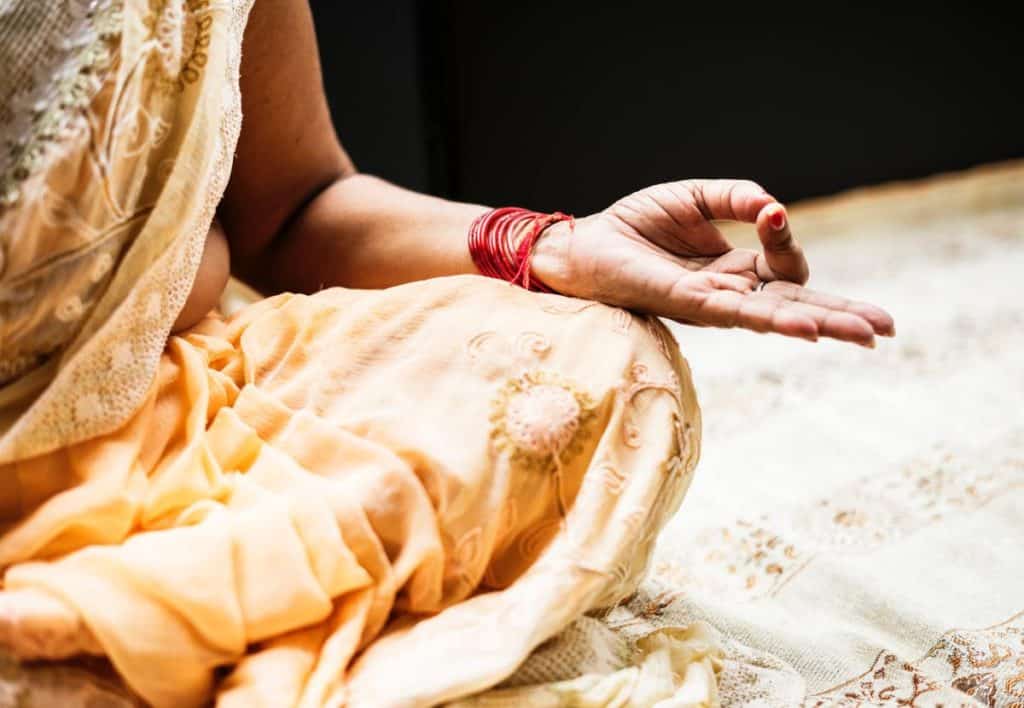 A person in a saree dress sitting in a meditation pose. Their fingers are folded into the Jnana Mudra hand pose.
