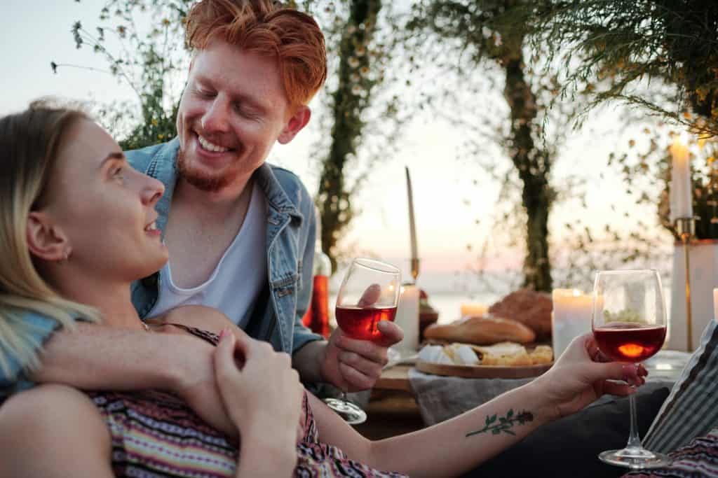 A male and female couple enjoys a picnic outdoors. The woman holds a glass of red wine.