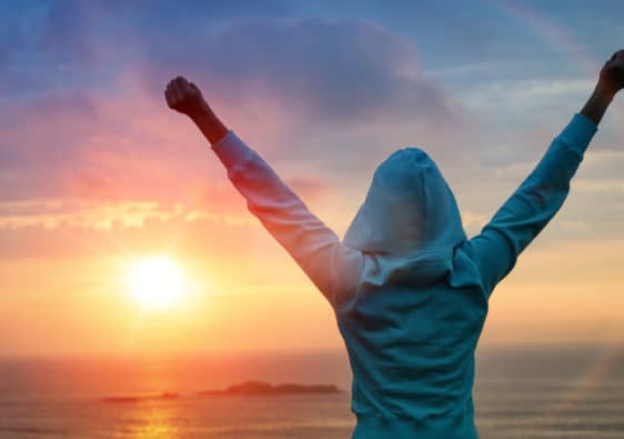 A person in a hoodie stands with their arms raised up in triumph as they look into the sunrise