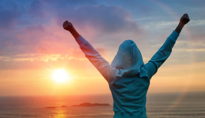 A person in a hoodie stands with their arms raised up in triumph as they look into the sunrise