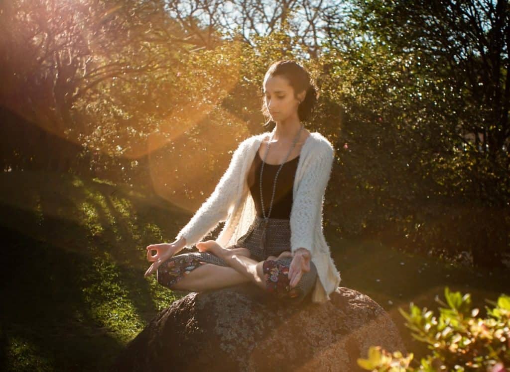 A woman sitting on a rock meditates outside near trees.