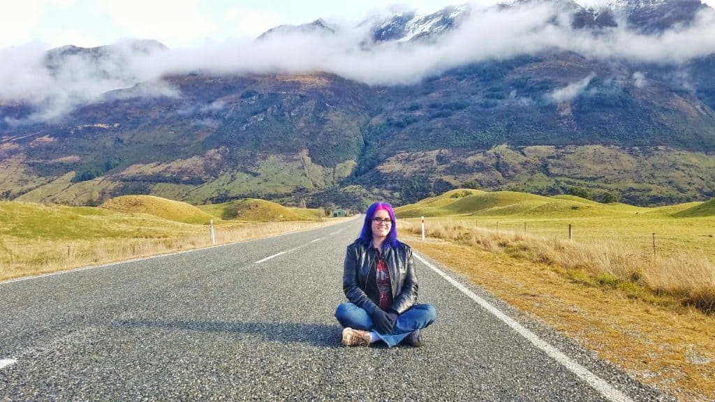 A woman with blue and pink hair sits cross legged in the street on a mountain.