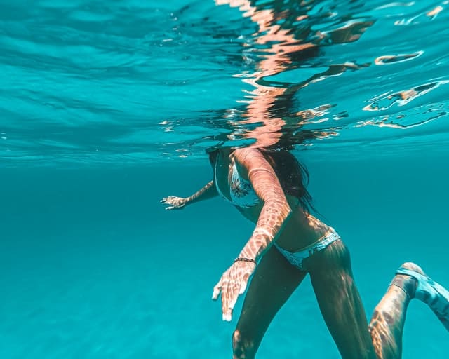 An underwater perspective of a woman in a bikini and flippers swimming in the ocean.