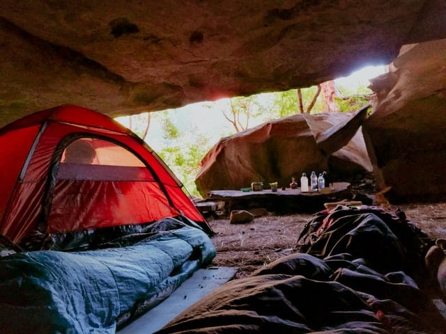 A red tent is pitched inside a cave underneath a giant red rock boulder. Sleeping bags, duffle bags, and water bottles are strewn about in the campsite.