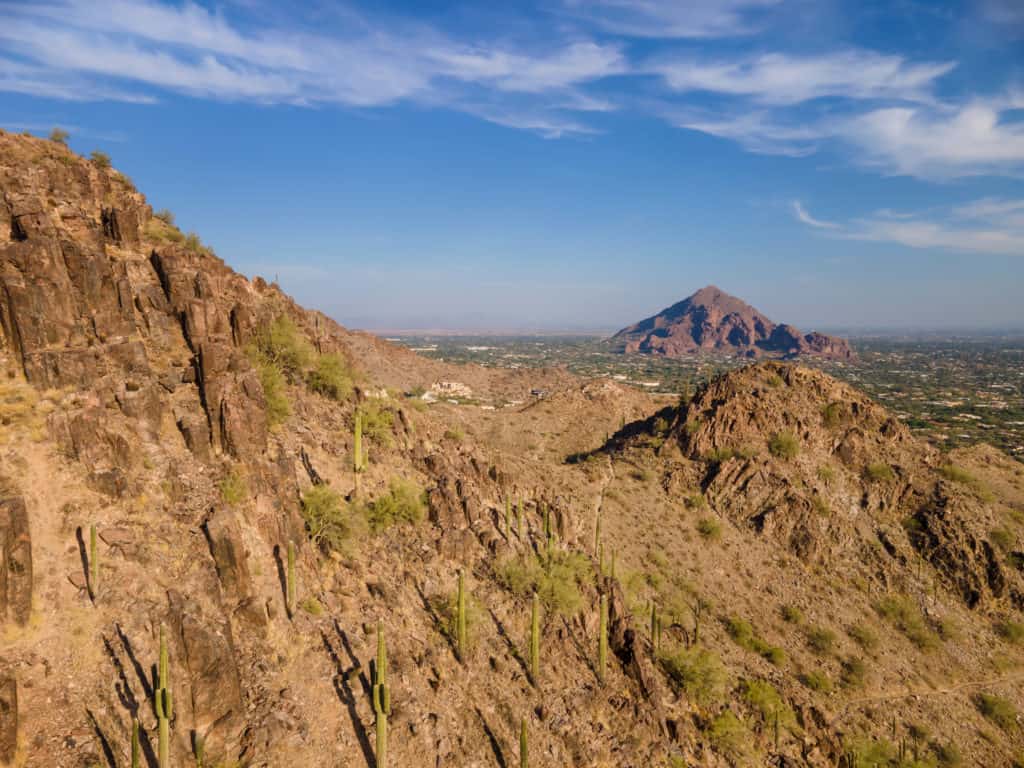 A small, desert mountain looms in the distance from the view of a hiking trail on another desert mountain.