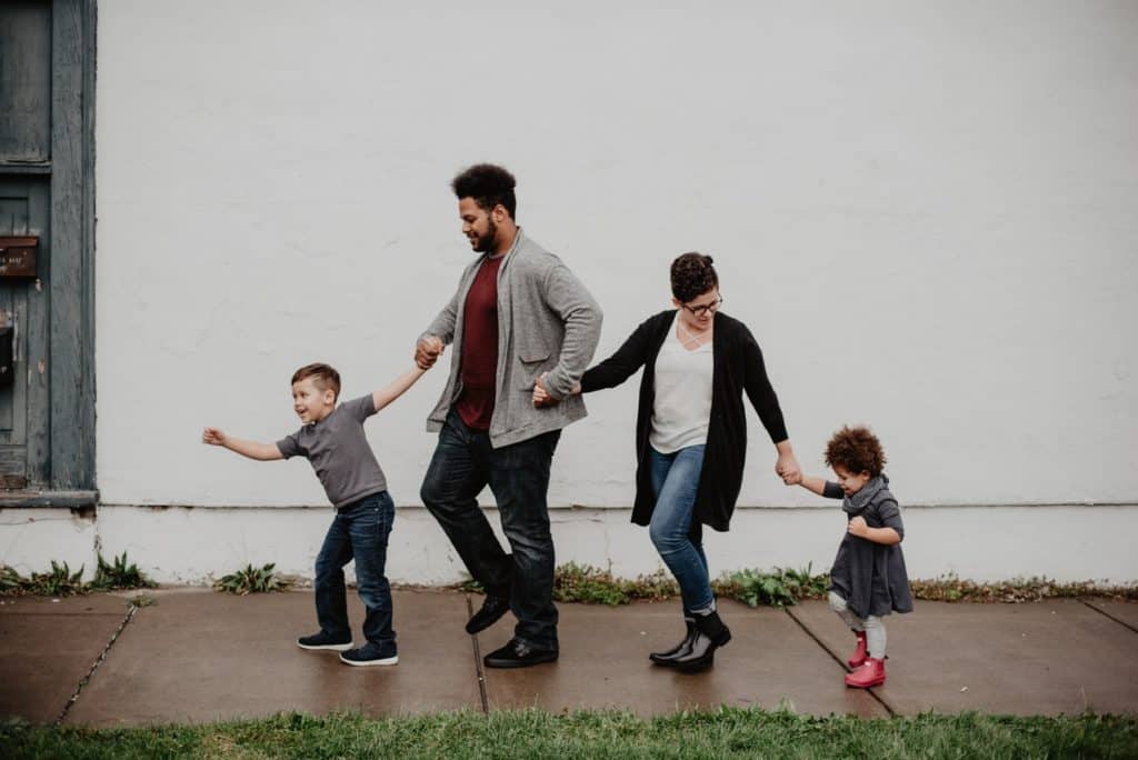 A biracial family of two parents and two young children walk on the sidewalk in the rain together, holding hands.
