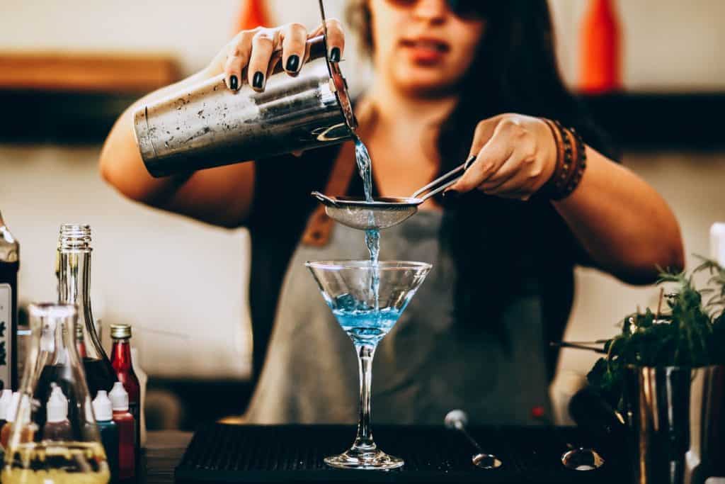 A woman pours a blue cocktail out of a shaker, through a strainer, and into a martini glass.