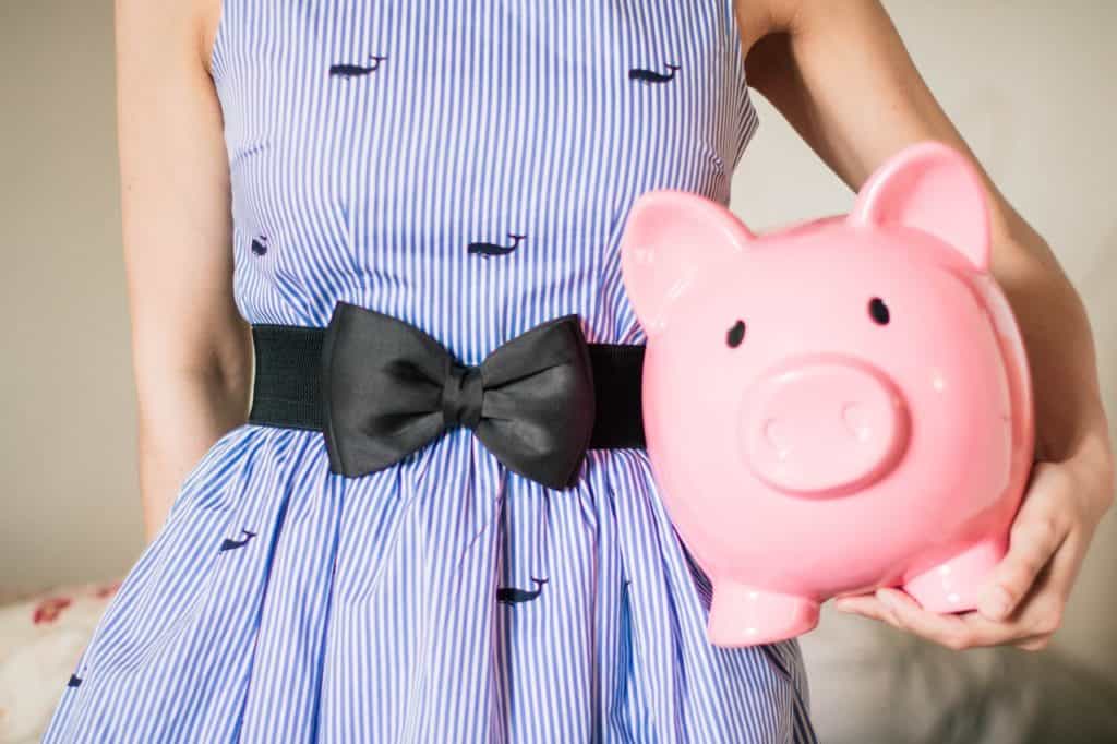 A woman in a blue dress holds an overly large pink piggy bank. 