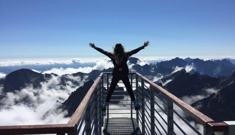 A woman stands on the railing of a look out at the top of a mountain. Her arms are thrown out in a power pose of triumph.