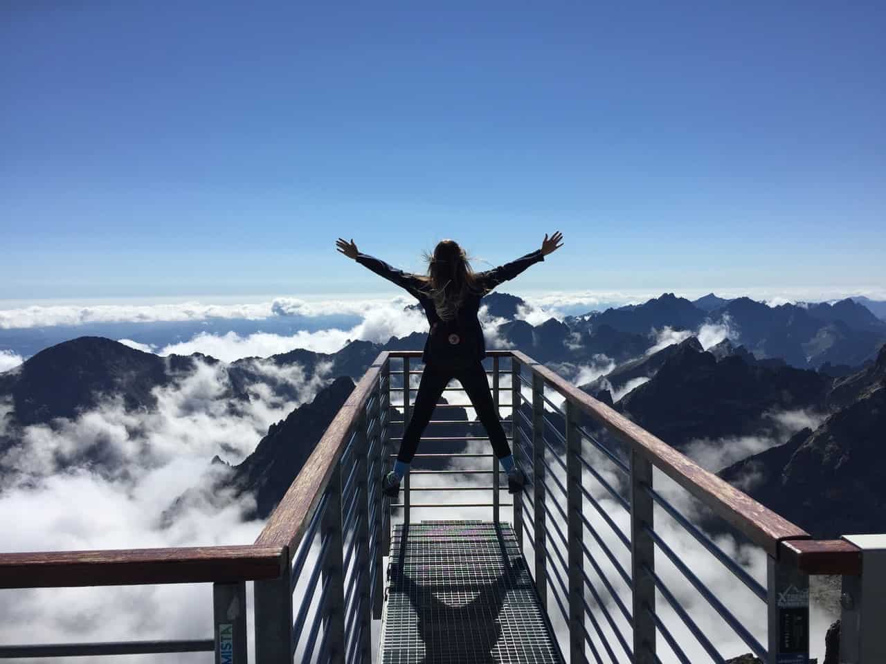 A woman stands on the railing of a look out at the top of a mountain. Her arms are thrown out in a power pose of triumph.