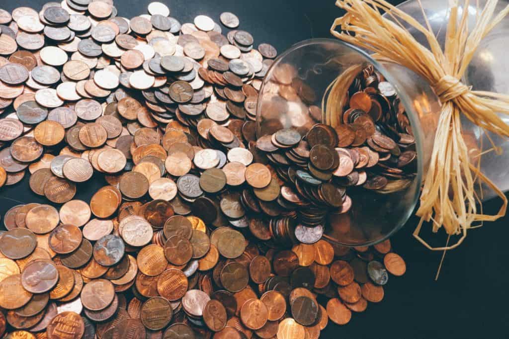 A clear, glass vase with a raffia bow is tipped over and an abundance of pennies have fallen out.