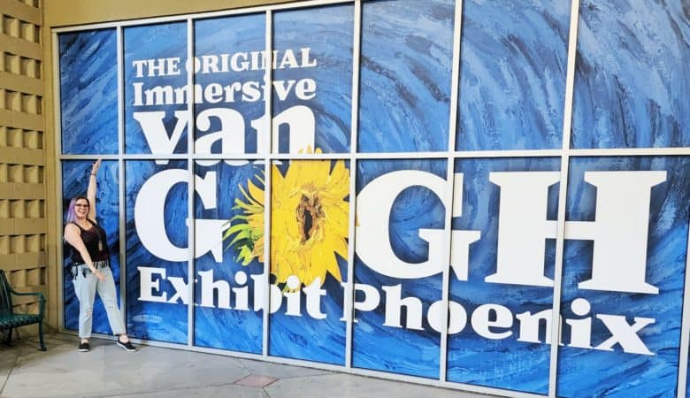 A woman poses in front of a sign that says, "The original immersive van gogh exhibit phoenix"