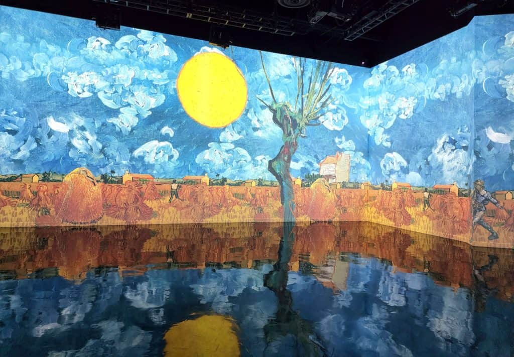 A Van Gogh painting of a yellow sun over wheat fields is projected on a tall wall and reflects off a glossy floor.