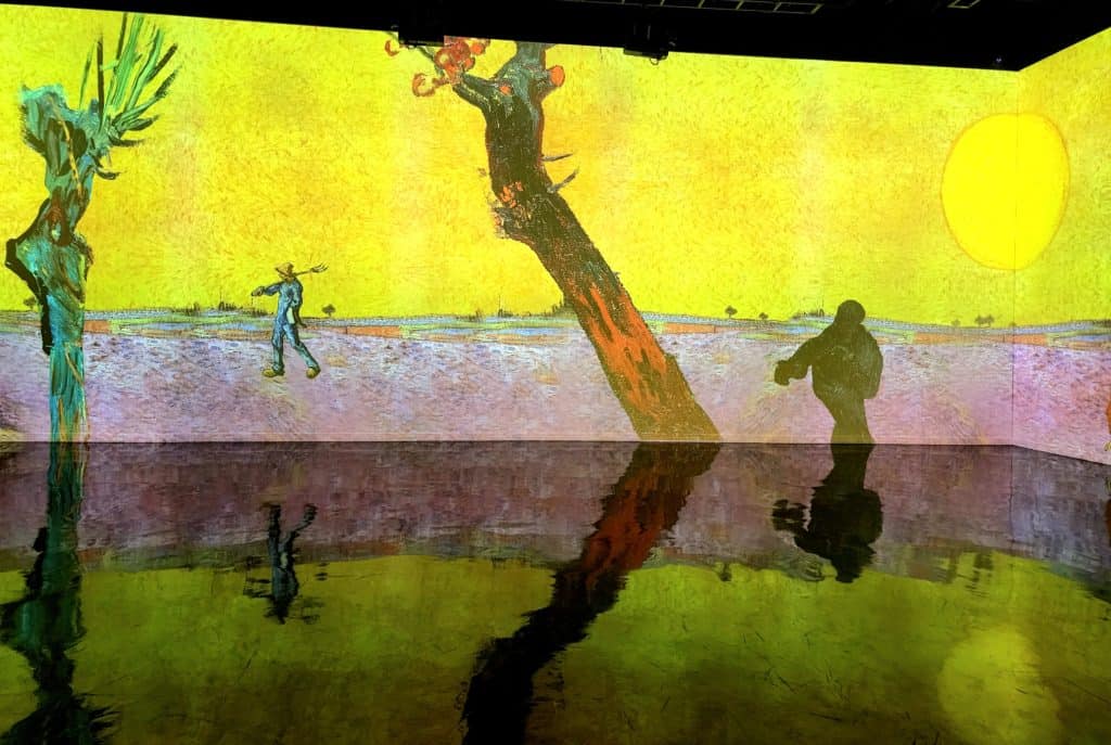 A Van Gogh painting of a yellow sun setting over an empty field is projected on a tall wall and reflects off a glossy floor.