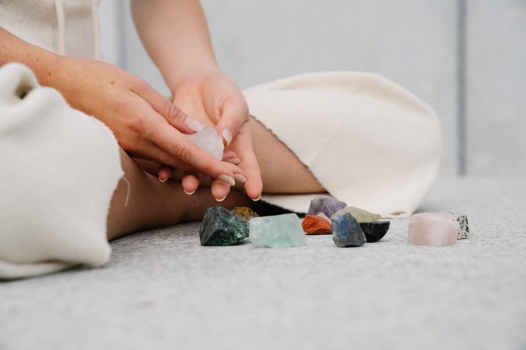 A person sits cross legged on the floor holding a clear quartz crystal while an assortment of other crystals are scattered next to them.