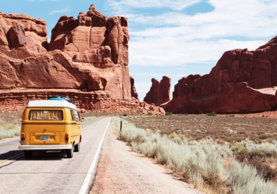A yellow Volkswagen bus drives on a desert road towards a horizon of red dirt mountains.