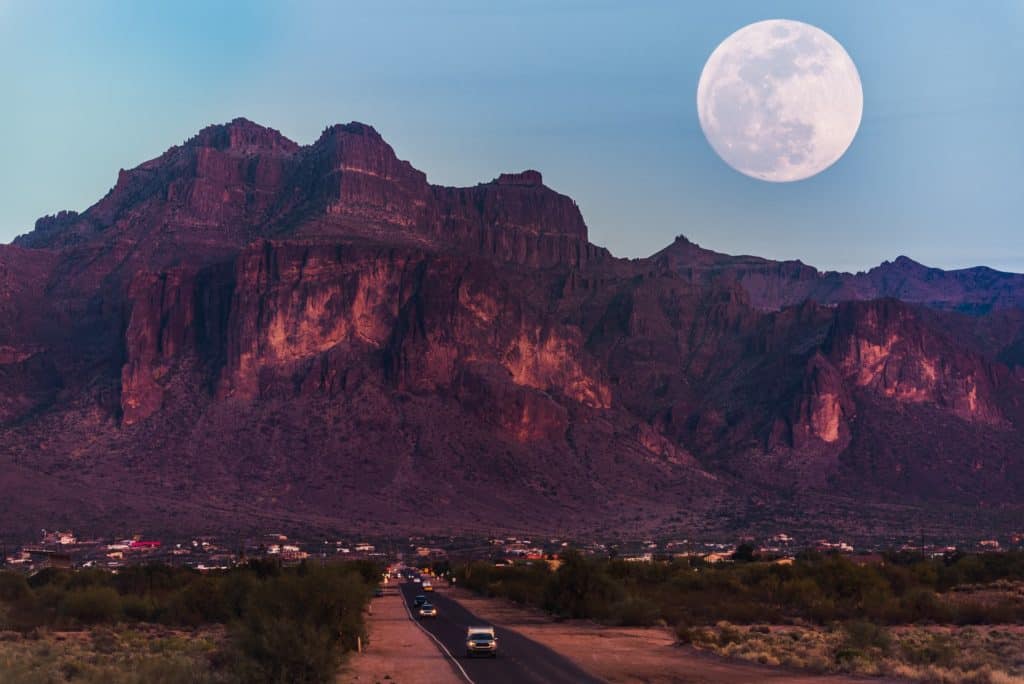 Dark red mountains on the edge of a small western town protrude up into the sky at dusk while a bright, full moon rises.