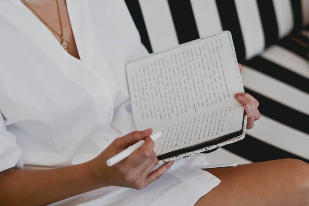 A woman sits on a couch and balances a journal on her left forearm while she writes in it.