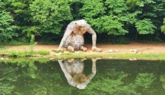 A giant wooden sculpture of a troll looks at its reflection in a pond