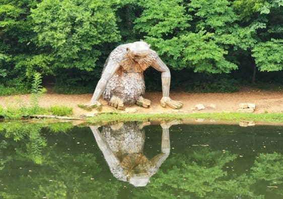 A giant wooden sculpture of a troll looks at its reflection in a pond