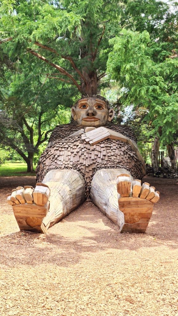 A giant wooden sculpture of a pregnant troll rests with her back against a tree trunk.