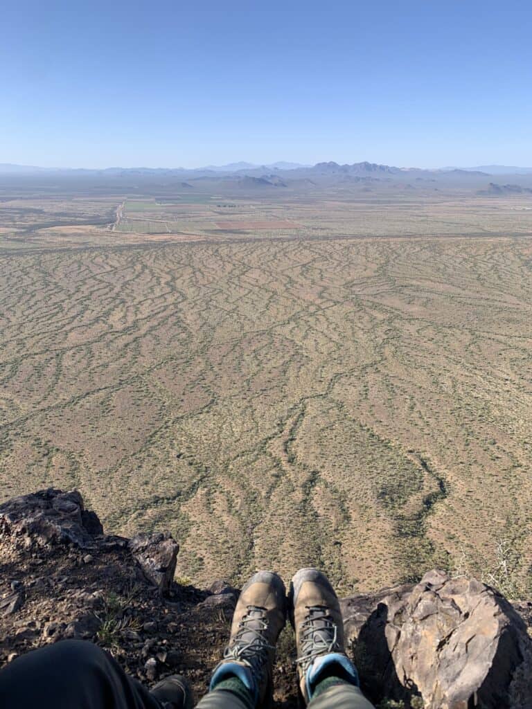 A pair of feet dangle over the edge of a cliff overlooking a desert landscape. Picacho Peak, one of the best peak hikes near Tucson. 