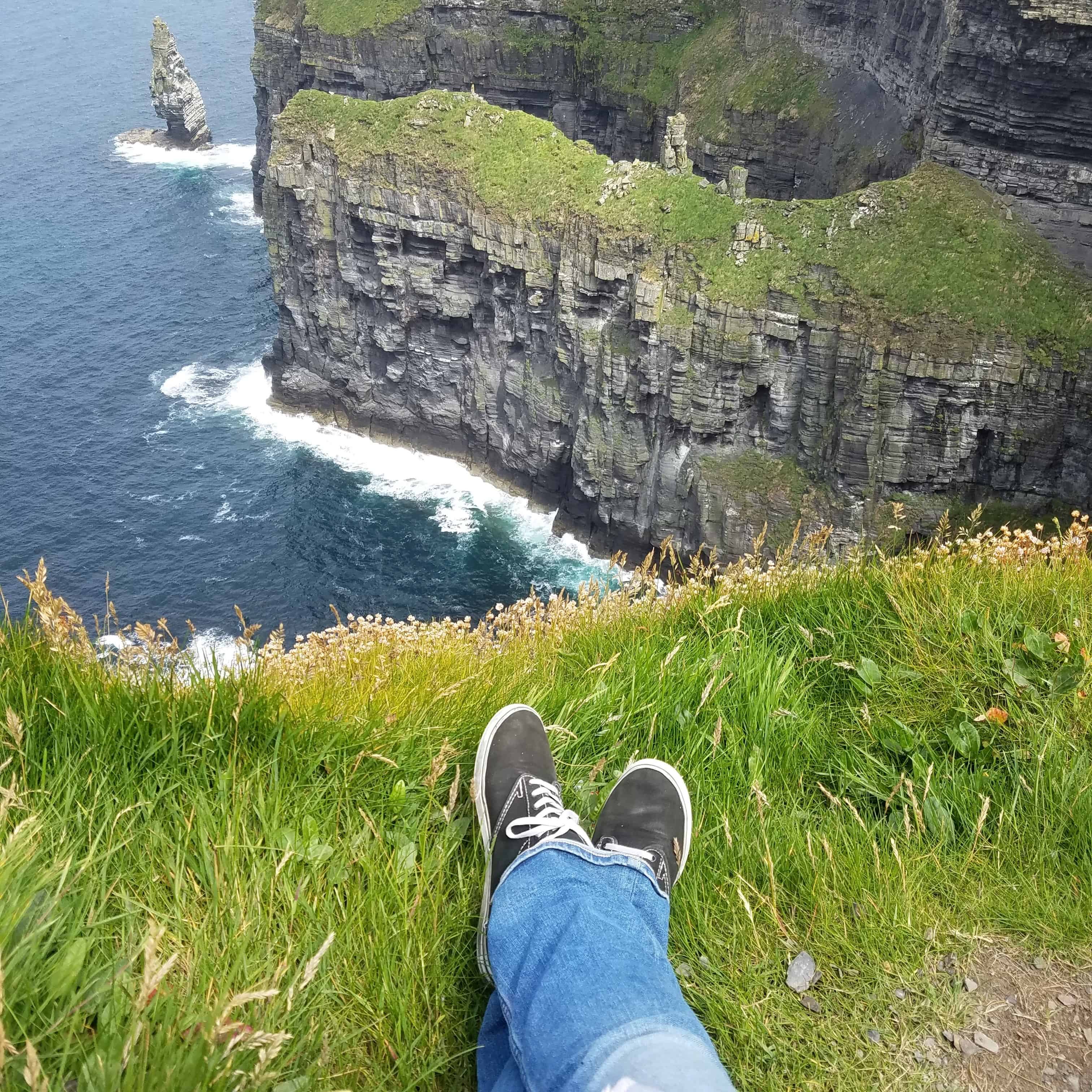 Legs wearing a beat up pair of vans and ripped jeans dangle over the edge of a cliff overlooking the ocean.
