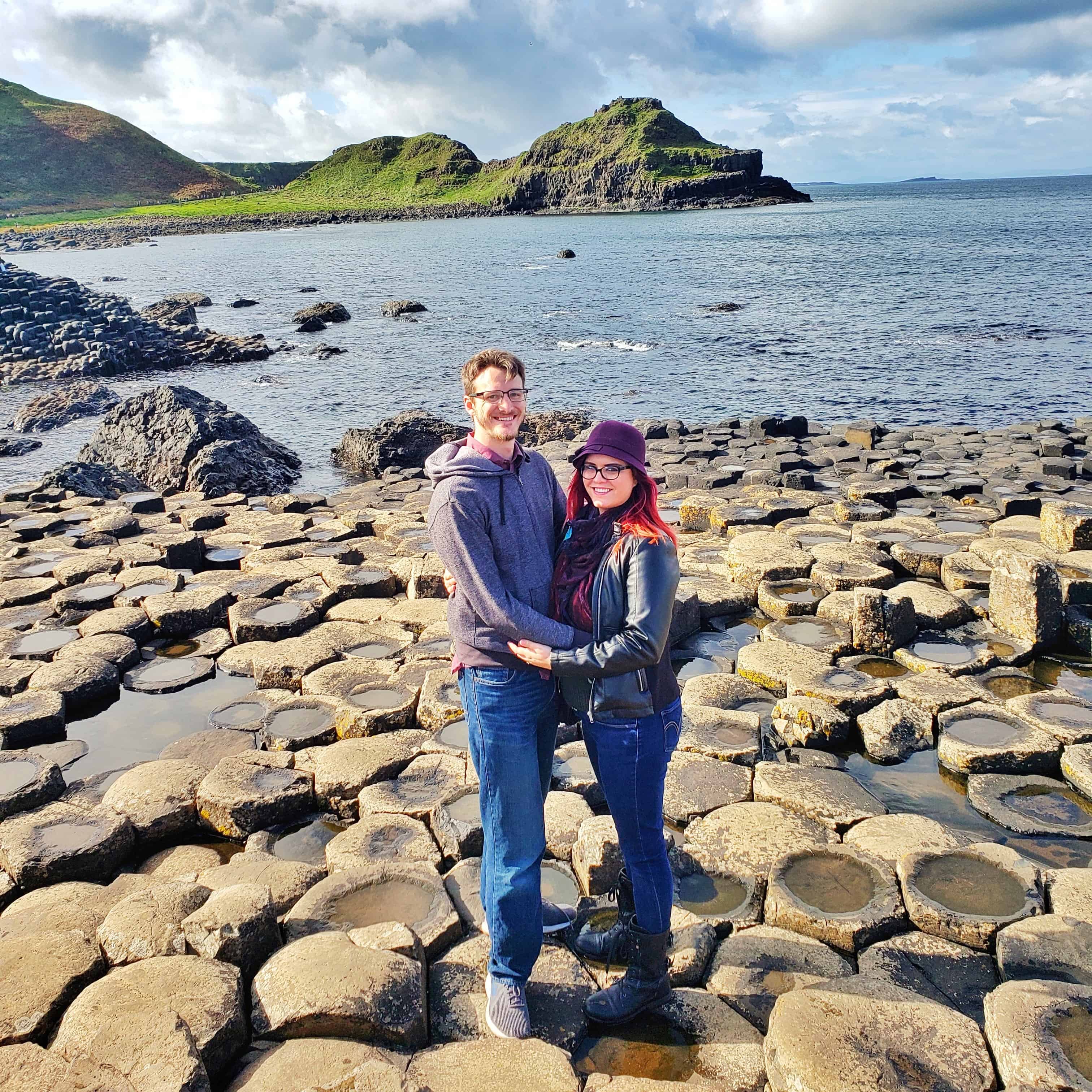 A couple (Mikey and Sckylar) stand on circular rocks next to the ocean.