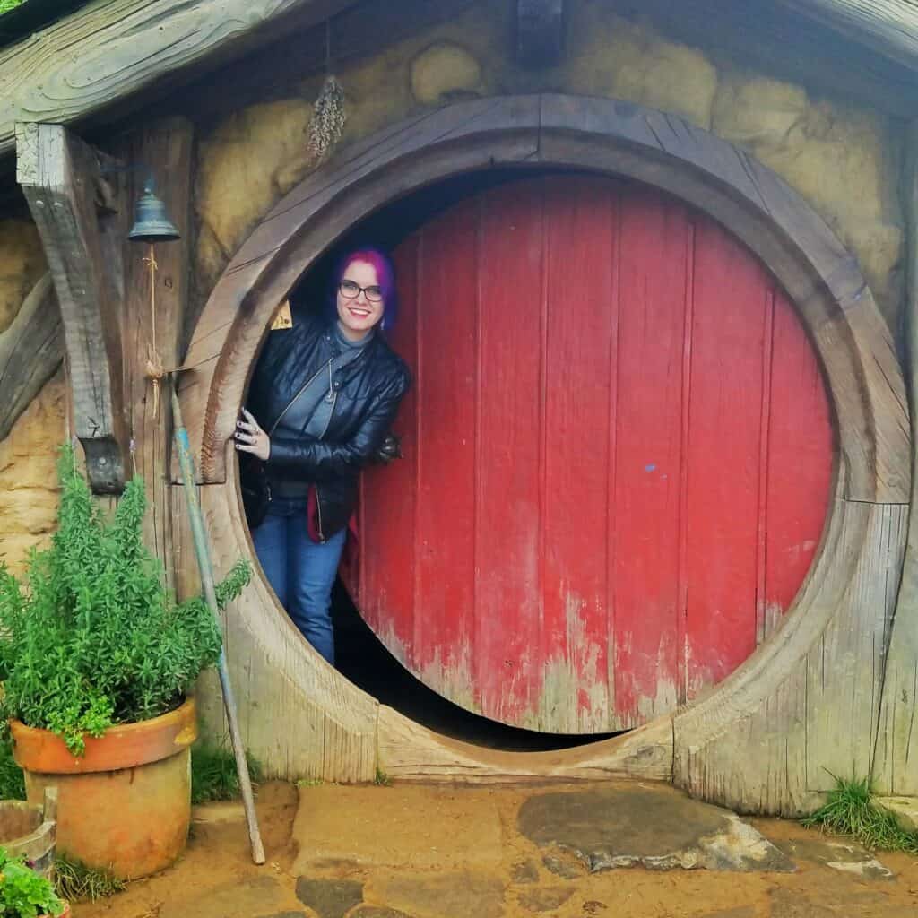 A woman with pink and blue hair (Sckylar) sticks her head out of a large red circular door, aka, a hobbit hole.