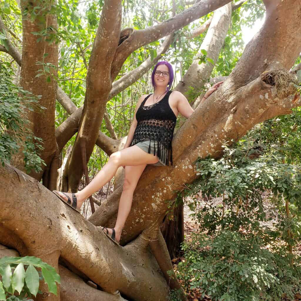 A woman with purple hair (Sckylar) stands on a limb of a large tropical tree.