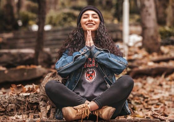 Woman sits cross-legged on dry leaves in woods with hands in namaste.