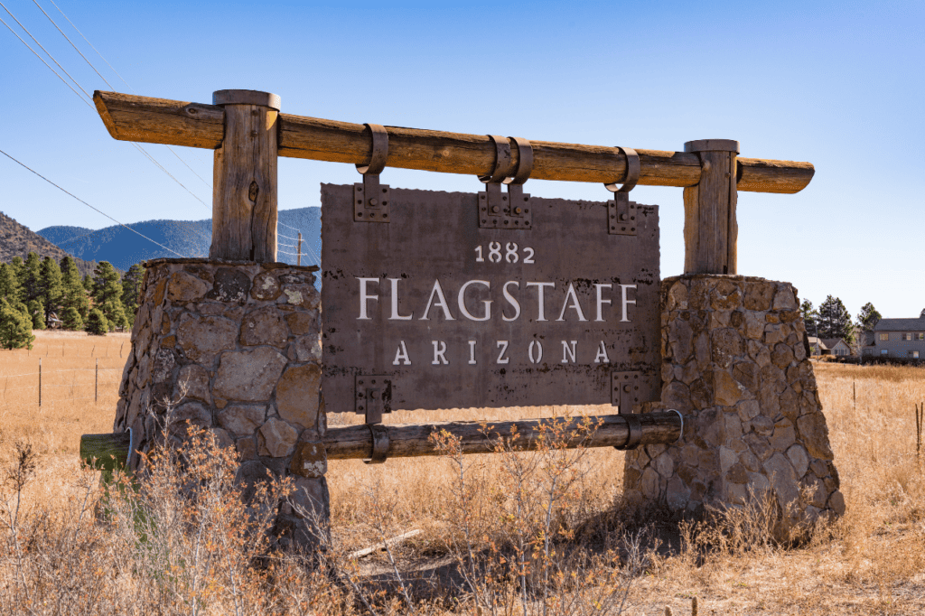 A large metal sign hanging from a wooden archway stuck inside two rock pedestals says "1882 Flagstaff Arizona."